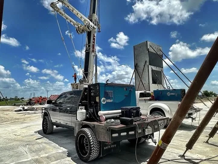 Black custom welding rig pickup with equipment in the flatbed beside cranes and industrial structures.