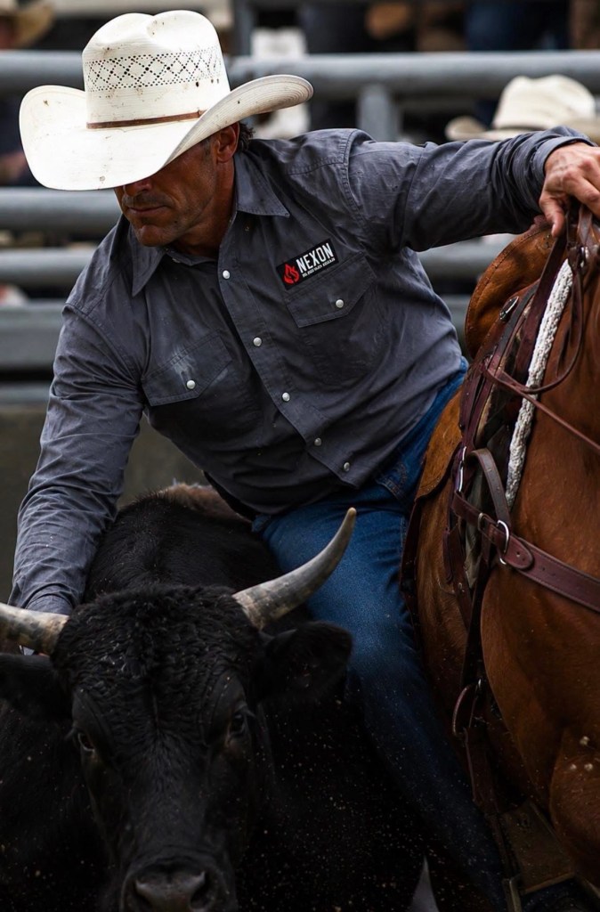 Cowboy in a white hat and gray NEXON pearl snap shirt leaning beside a black bull and saddled horse.